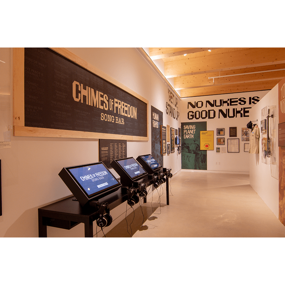 An exhibit titled "Chimes of Freedom Song Bar," featuring a wall with song titles, a row of interactive touch-screen kiosks with headphones, and surrounding wall displays with protest-themed posters and artifacts.