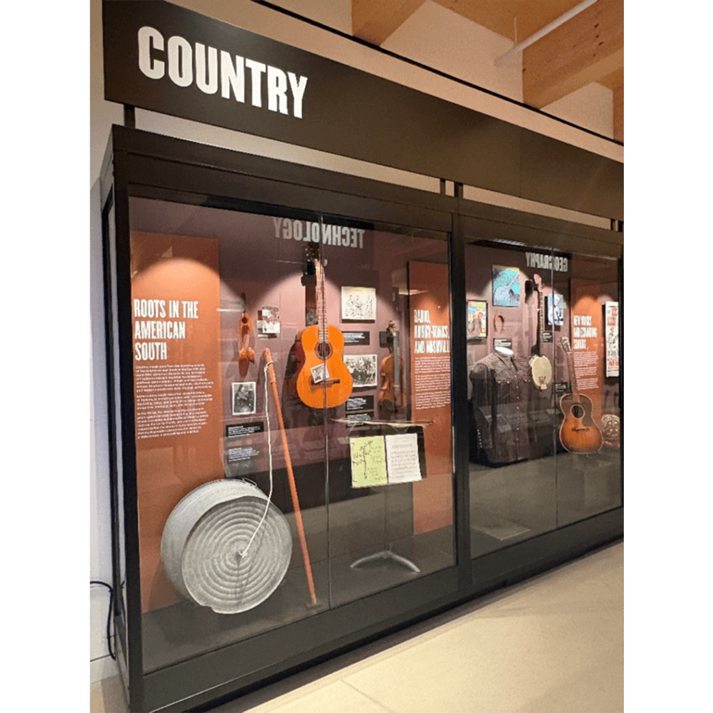 Museum exhibit themed "Country." Displays include a guitar, washboard, banjo, and various photos, highlighting the music's Southern roots. Warm lighting enhances a nostalgic tone.