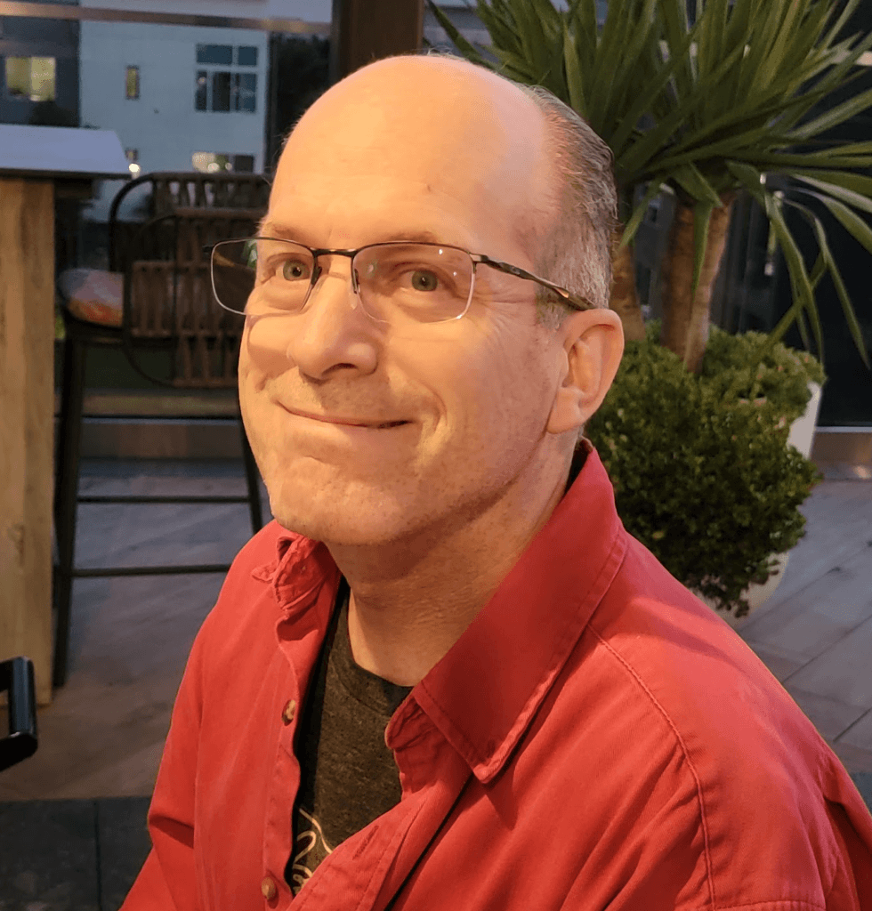 A portrait of Dan Loughran, a fair-skinned man with short hair and glasses, wearing a bright red button-down shirt over a dark t-shirt.
