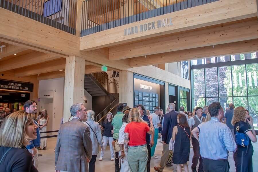 A photograph of a bustling event in the light-filled, wood-beamed interior of the Bruce Springsteen Center for American Music. A large group of people are socializing in the main hall, with a sign reading "Hard Rock Hall" visible on a prominent overhead beam, and signage for the "Jon Landau American Music Gallery" on the left.