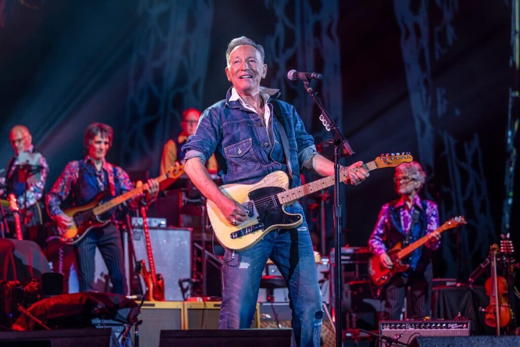 Bruce Springsteen passionately plays guitar onstage, wearing a denim jacket. Behind him, a band in vibrant shirts plays instruments against a backdrop of dramatic lighting.