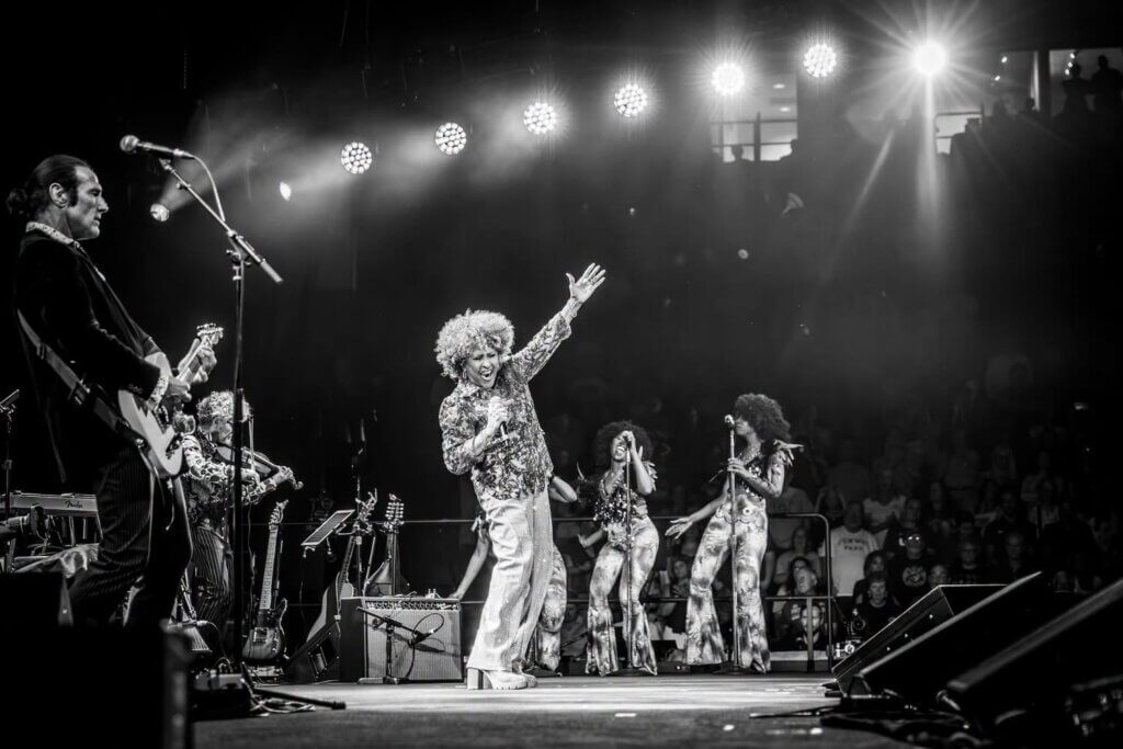 Black and white image of a lively concert scene. Darlene Love waves on stage, with musicians and singers in sparkling outfits under bright lights.