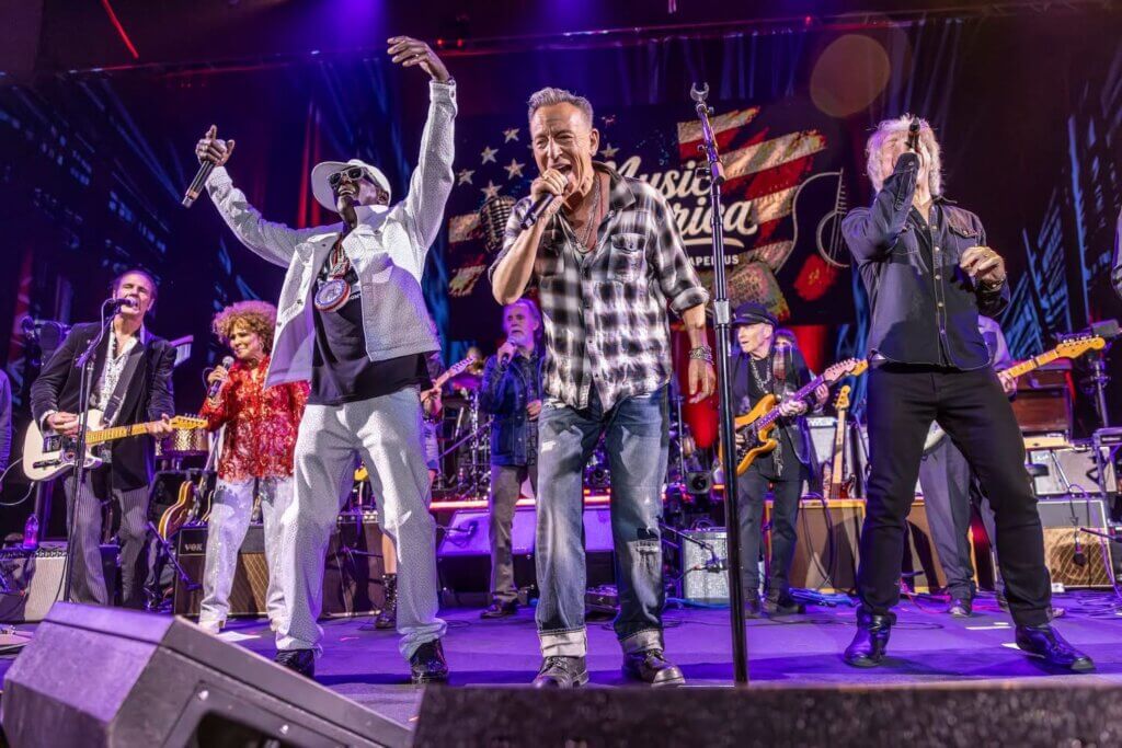 A lively concert scene with seven performers on stage, singing and playing guitars. The crowd is energized, with colorful lighting and vibrant American-themed backdrop. Flavor Flav, Bruce Springsteen and Jon Bon Jovi across the front of the stage.