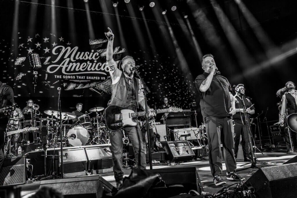 Black and white photo of a lively band performing on stage under bright lights. One musician raises a hand; a backdrop reads "Music of America." Energetic atmosphere. Bruce Springsteen and Ken Casey across the front of the stage.