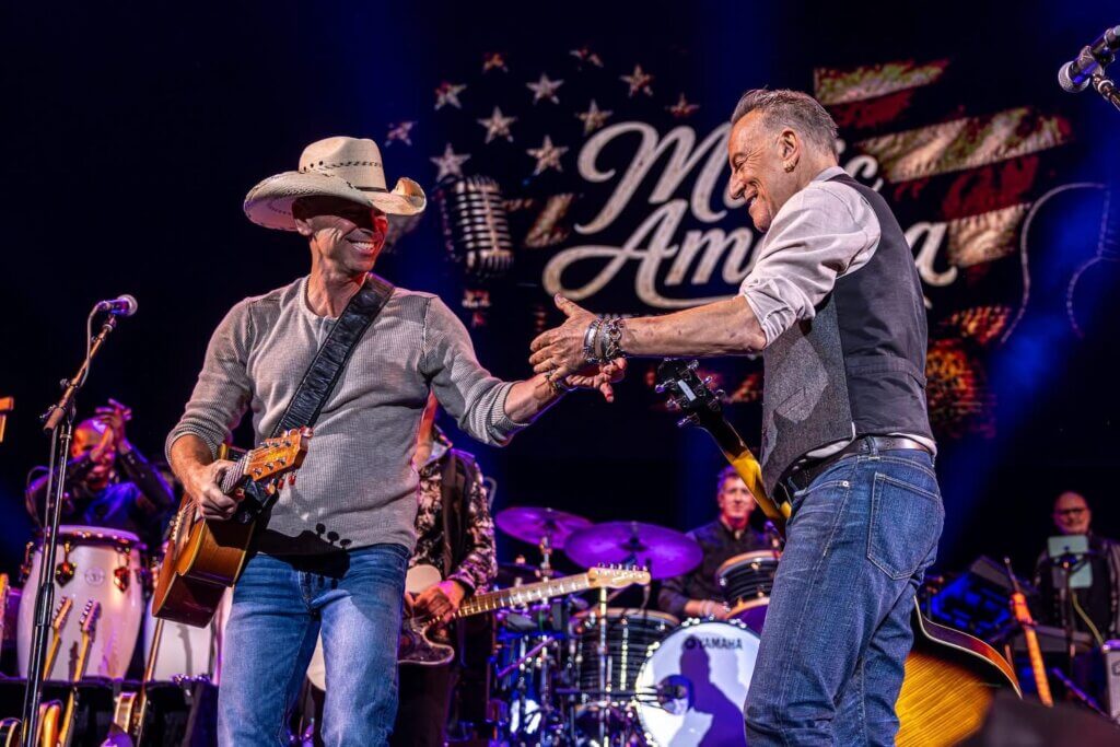 Kenny Chesney and Bruce Springsteen on stage share a friendly handshake, exuding joy. One wears a cowboy hat, holding a guitar. "Music America" is displayed behind them.