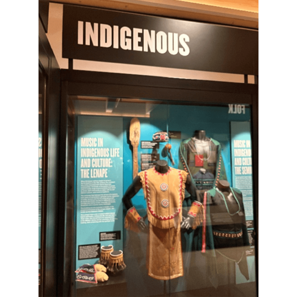 Exhibit display titled "Indigenous" showcasing traditional Lenape clothing. Includes a beaded dress, drum, and cultural artifacts. The setting is educational and respectful.