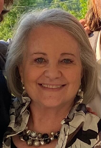 A portrait of a woman with shoulder-length grey hair, smiling warmly at the camera. She is wearing a patterned top with a ruffled collar and a large, beaded statement necklace.