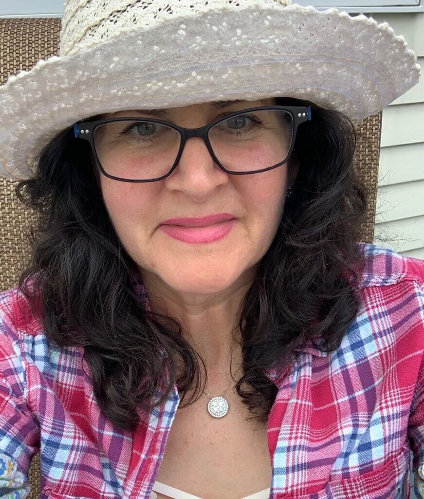 A selfie portrait of a woman with dark wavy hair wearing a straw sun hat, black-framed glasses, and a red and blue plaid shirt. She is wearing a delicate necklace with a circular pendant and has bright pink lipstick on.