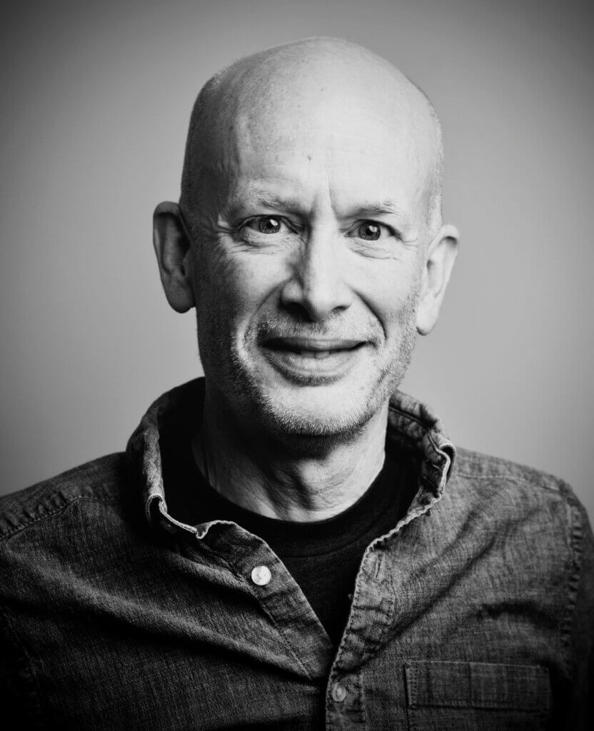 A black-and-white headshot portrait of a bald, middle-aged man smiling warmly at the camera, wearing a dark t-shirt under an unbuttoned denim shirt.