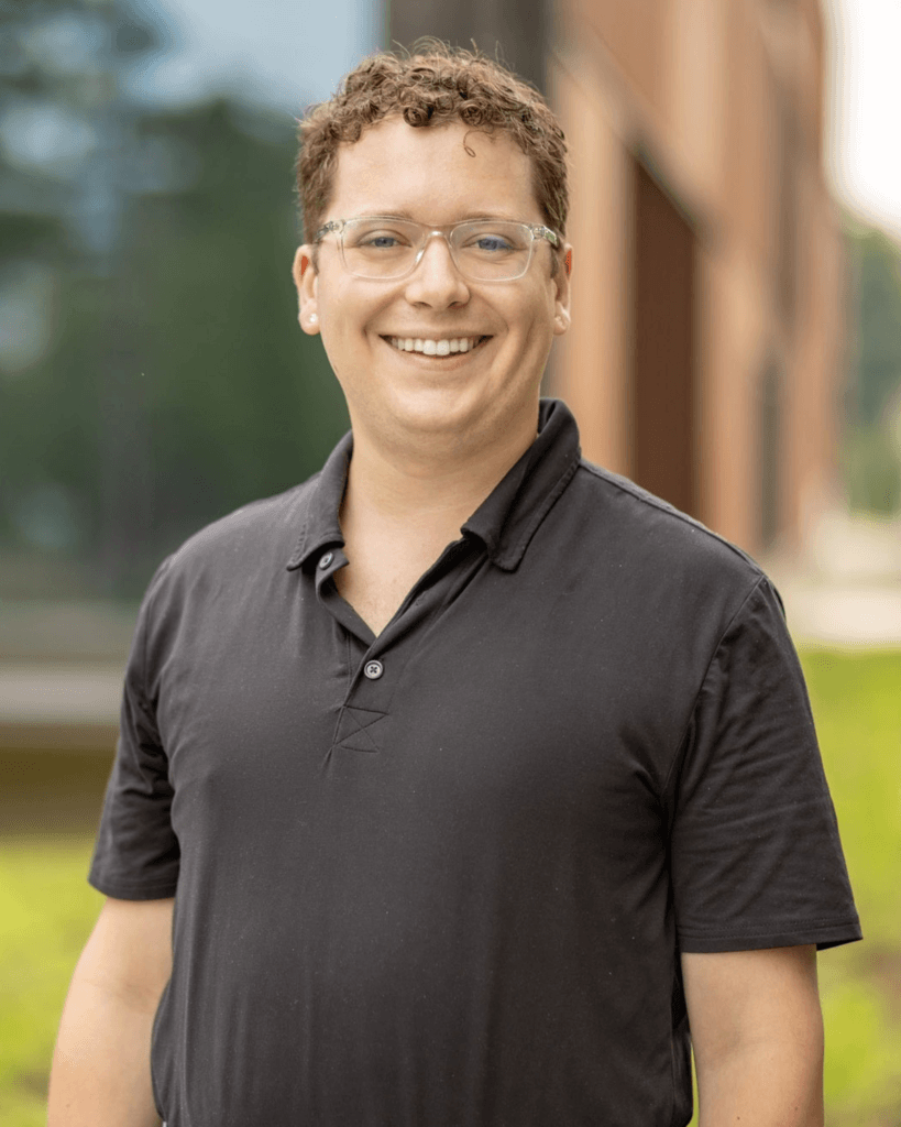 Portrait of a smiling person with short curly hair and clear-framed glasses wearing a black polo shirt, standing outdoors with a blurred building in the background.