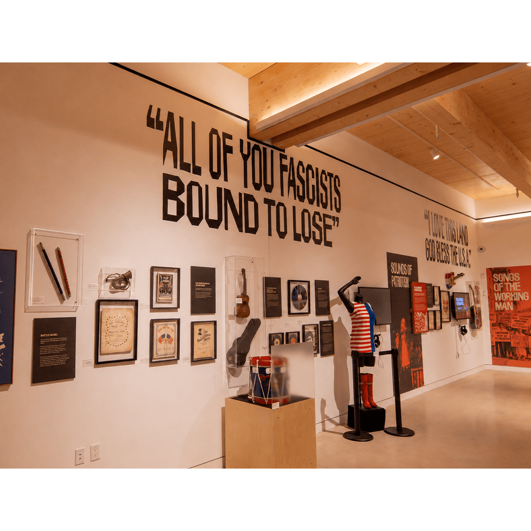 A view inside the Bruce Springsteen Center for American Music featuring the 'Chimes of Freedom' exhibit. The wall prominently displays the lyrics 'All of you fascists bound to lose' in large, black lettering, with various historical music artifacts, framed posters, a vintage drum, and a mannequin wearing a patriotic-themed outfit displayed below. Further along the wall, additional signage reads 'I love this land, God bless the U.S.A.' and 'Songs of the working man