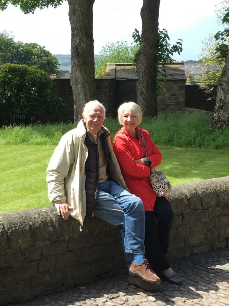 A smiling senior man in a cream jacket and blue jeans sits next to a smiling woman in a bright red jacket on a stone wall in a grassy garden setting.
