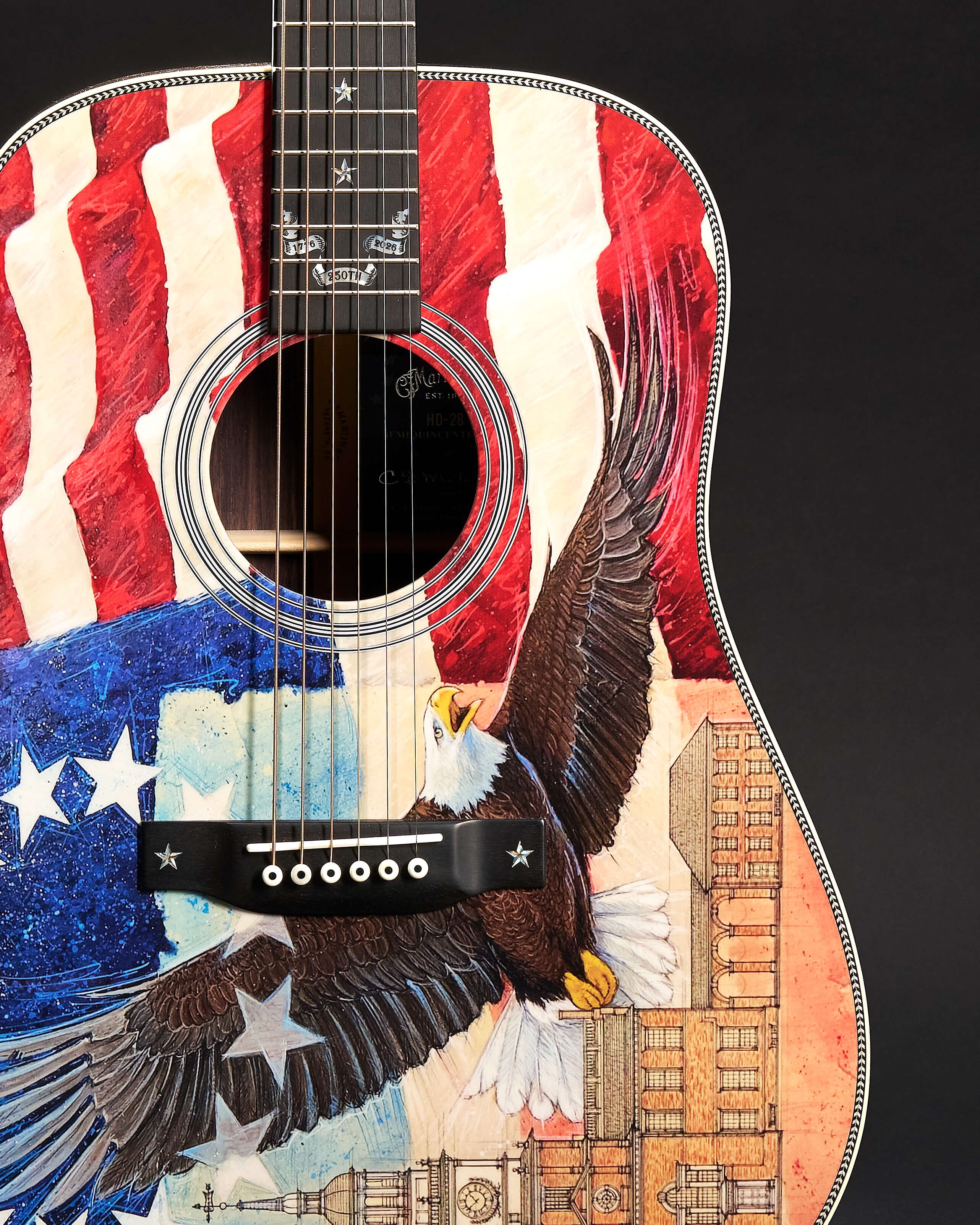 Close-up of a Martin HD-28 Semiquincentennial acoustic guitar body. The top is painted with a vibrant patriotic theme, featuring a waving American flag, a soaring bald eagle with outstretched wings, and intricate line drawings of historical architecture. The fretboard includes custom star inlays and a "250th" banner.