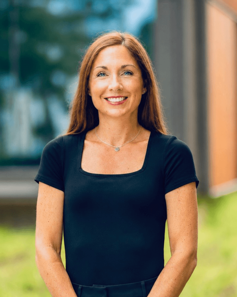 Portrait of a smiling person with long brown hair wearing a black short-sleeve top and a silver necklace, standing outdoors with a blurred building in the background.