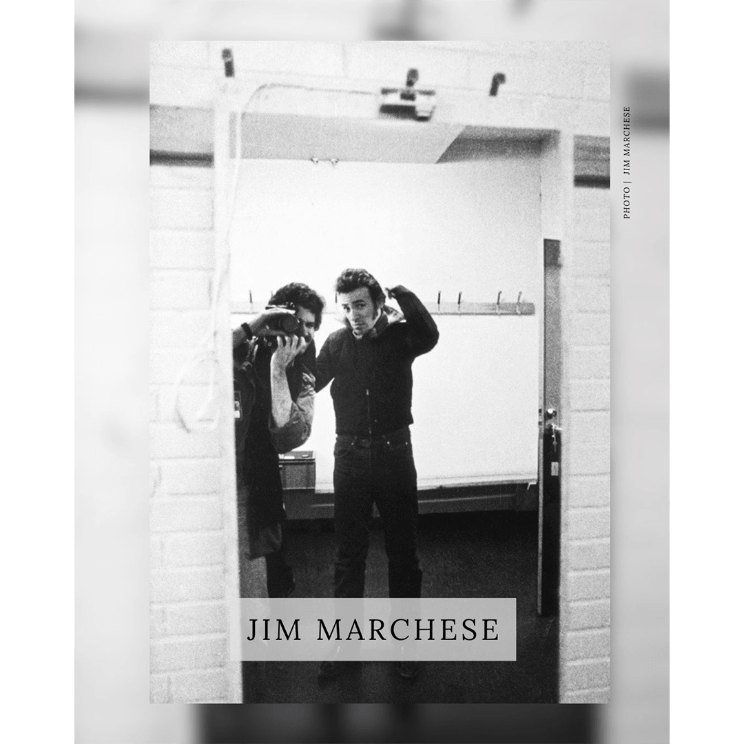 A black-and-white photograph by Jim Marchese showing a reflection of Bruce Springsteen in a mirror. Springsteen is seen adjusting his collar, while Marchese is visible in the reflection as he takes the photograph with his camera. The name 'JIM MARCHESE' is overlaid in a box at the bottom of the image