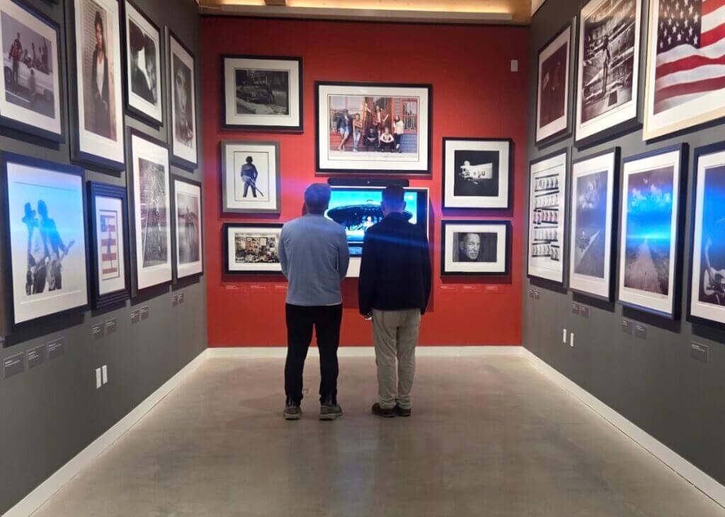 Two visitors viewed from behind standing in the center of a photography exhibit, surrounded by framed black-and-white and color photos on dark gray and red accent walls.