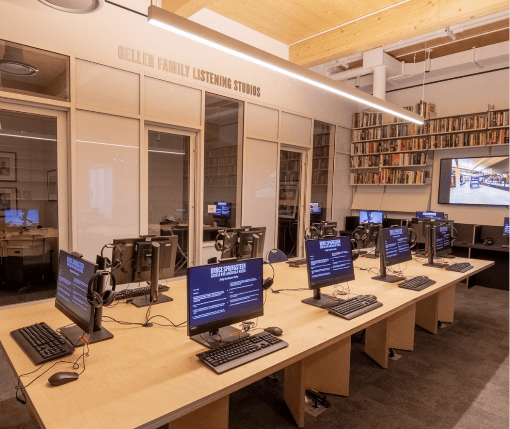 The Geller Family Listening Studios at the Bruce Springsteen Center for American Music, featuring a long wooden table with multiple computer workstations, headphones, and a large bookshelf lining the wall.