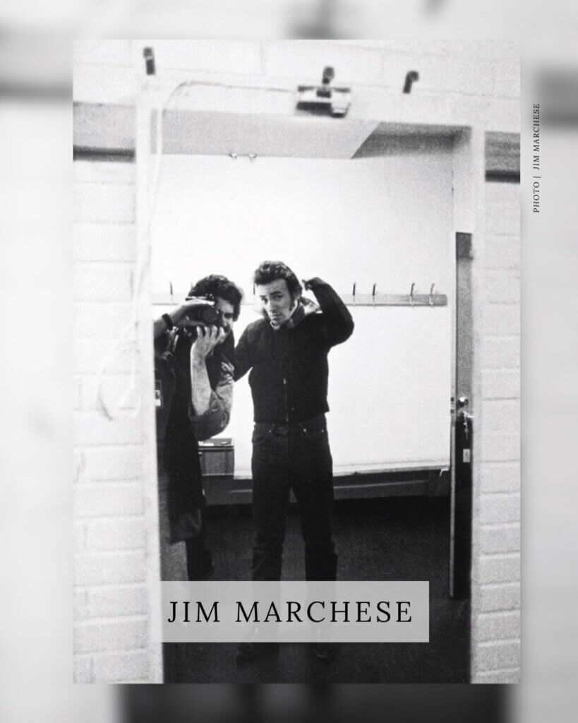 A black-and-white photograph by Jim Marchese showing a reflection of Bruce Springsteen in a mirror. Springsteen is seen adjusting his collar, while Marchese is visible in the reflection as he takes the photograph with his camera. The name 'JIM MARCHESE' is overlaid in a box at the bottom of the image