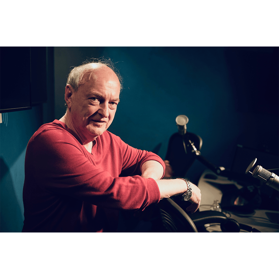 Man seated at a SiriusXM microphone in a recording studio, wearing a red sweater and looking toward the camera.