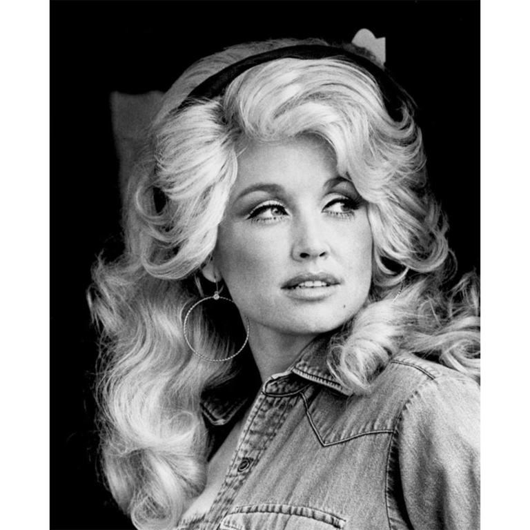 Black and white portrait of a young Dolly Parton with voluminous blonde hair, hoop earrings, and a denim shirt, looking off to the side.
