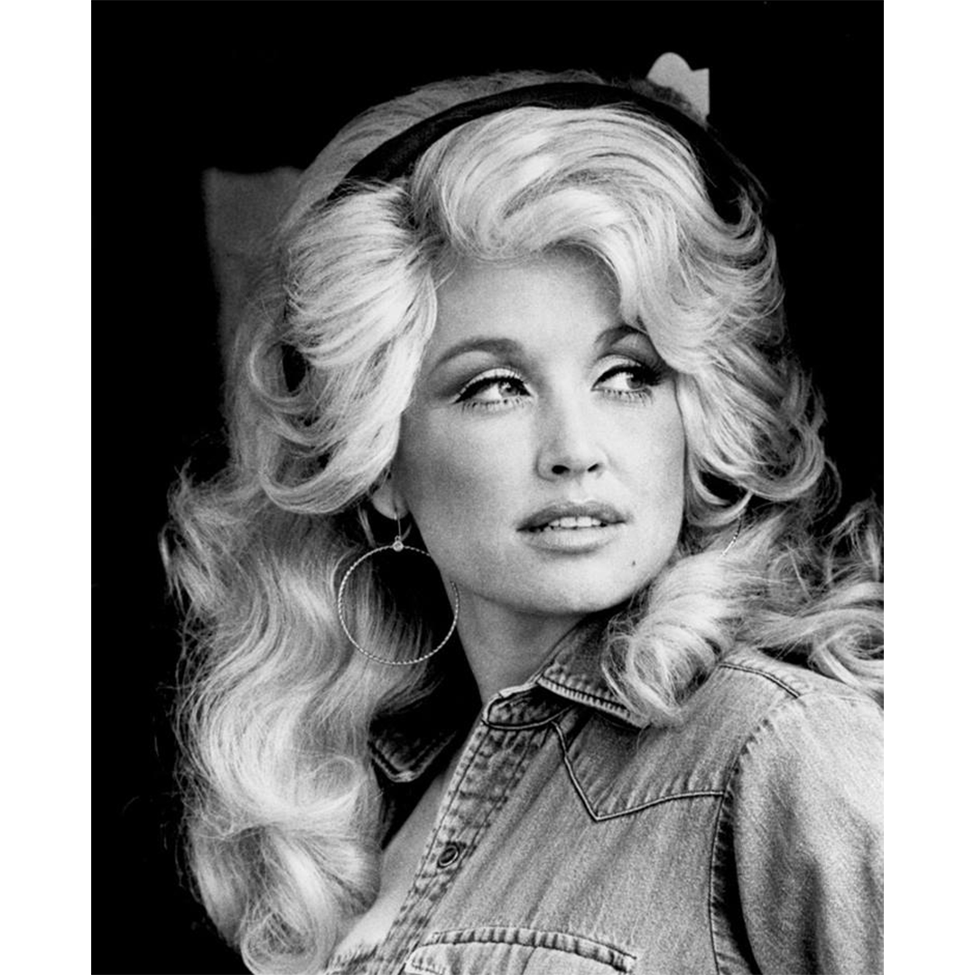 Black and white portrait of a young Dolly Parton with voluminous blonde hair, hoop earrings, and a denim shirt, looking off to the side.
