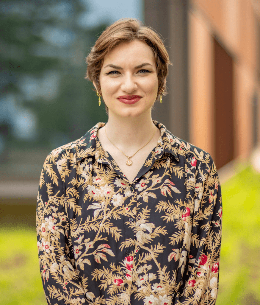 A woman with short light-brown hair stands outdoors, facing the camera and smiling. She wears a dark floral-patterned button-up blouse, gold drop earrings, and layered gold necklaces. The background is softly blurred with greenery and a warm-toned building.