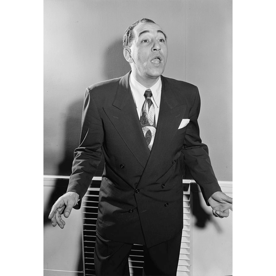 Black and white portrait of American musician and entertainer Louis Prima in a double-breasted suit and patterned tie, gesturing with his hands wide while singing.