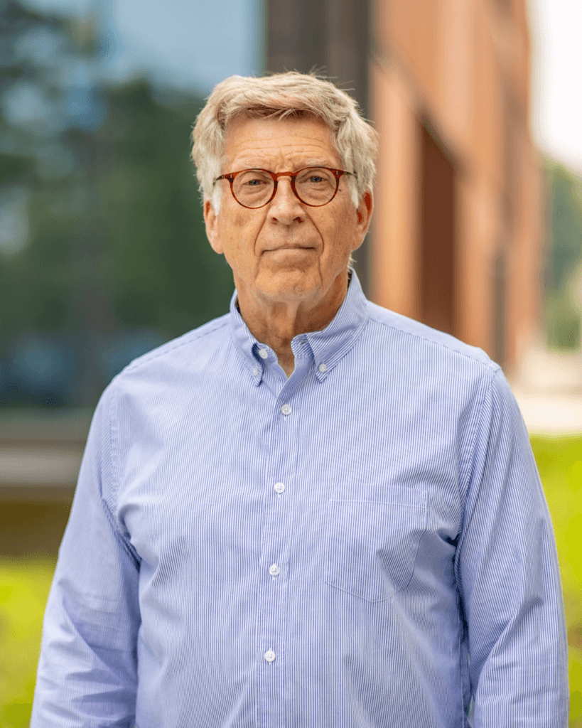 An older man - Bob Santelli - with gray hair and glasses stands outdoors in front of a softly blurred building and greenery, wearing a light blue button-down shirt.