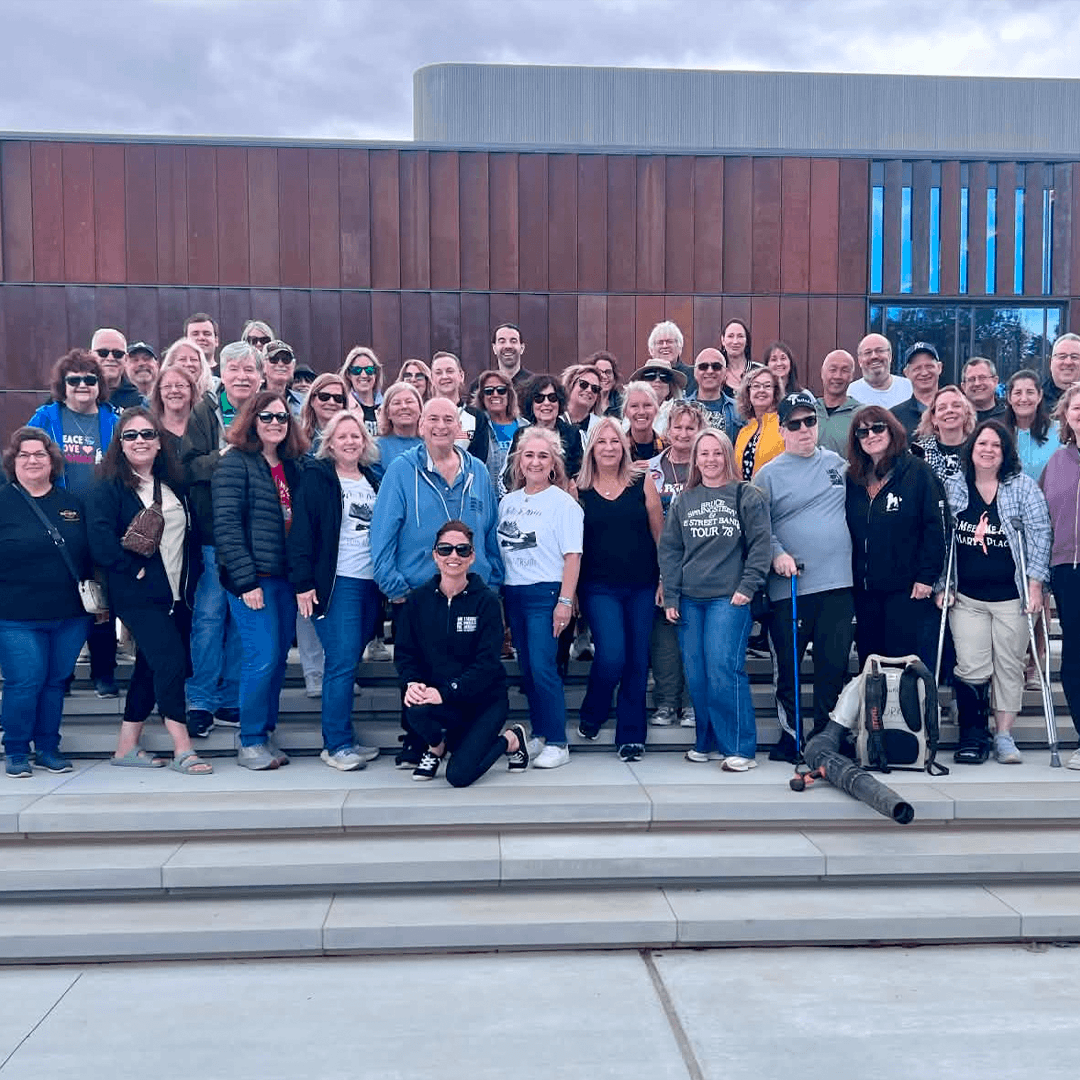 A large group of adults pose together on wide outdoor steps in front of a modern building with a curved silver roof and wood-paneled facade. Several people wear sunglasses, jackets, or event T-shirts, and a few mobility aids are visible. The sky is overcast.