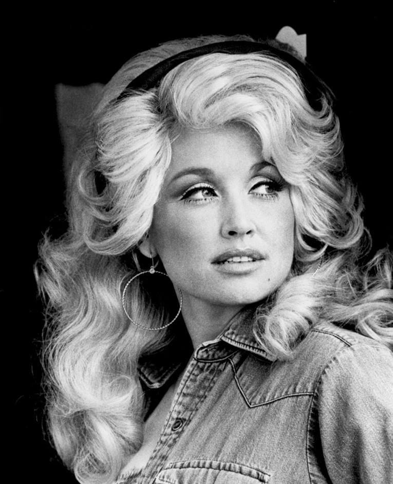 Black and white portrait of a young Dolly Parton with voluminous blonde hair, hoop earrings, and a denim shirt, looking off to the side.