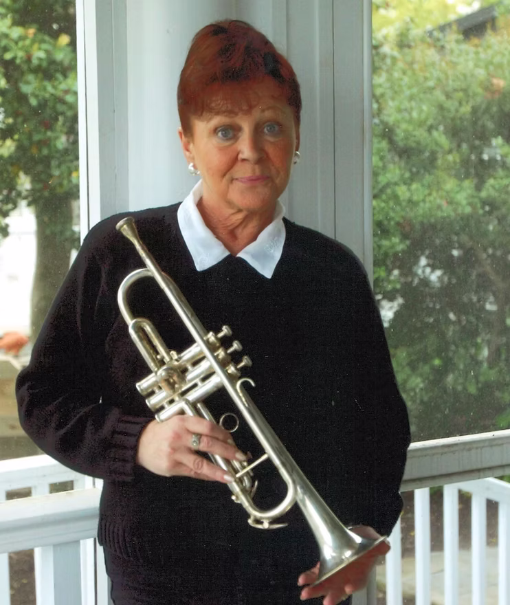 Portrait of a woman with red hair wearing a black sweater and white collared shirt, holding a silver trumpet on a porch.