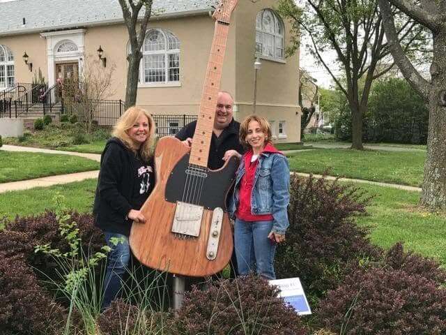 Three adults pose outdoors beside a large wooden guitar sculpture in a landscaped area. A tan building with tall arched windows, trees, sidewalks, and lawns is visible in the background.