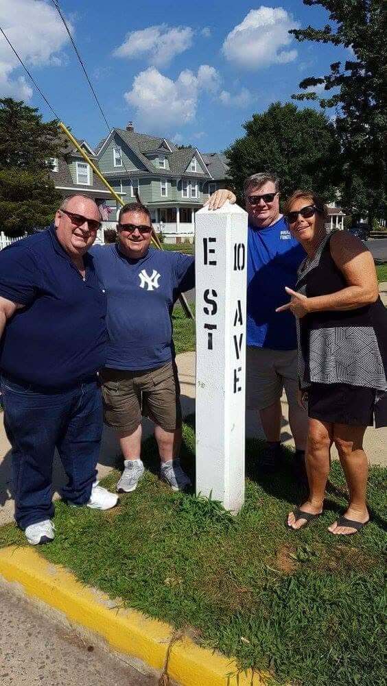 Four adults pose beside a white roadside marker reading “EAST” and “AVE” in a residential neighborhood. Three men and one woman are standing on a grassy verge near a yellow-painted curb, with large older homes, trees, utility wires, and a bright blue sky in the background.