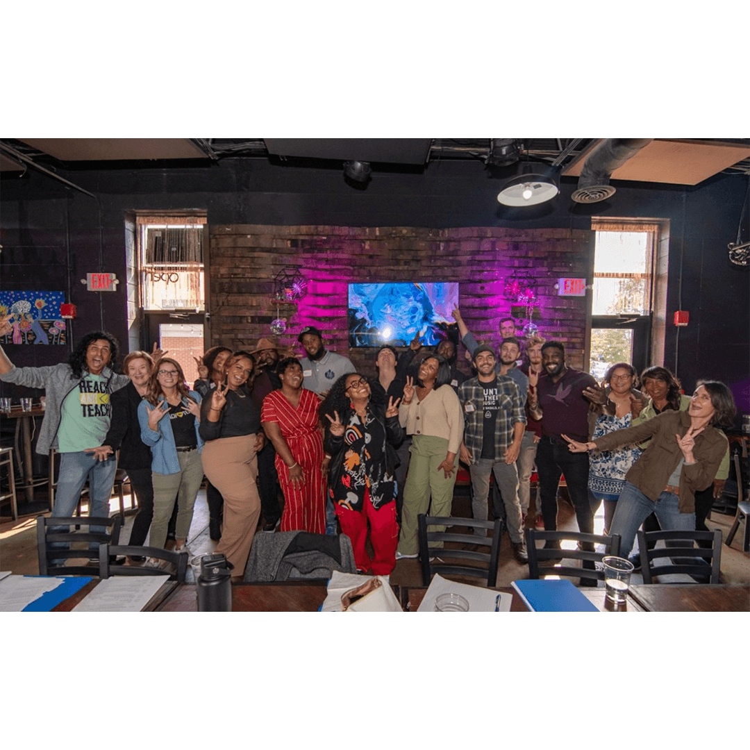 Group photo of people gathered inside an event space with a wooden accent wall, posing and smiling with tables and chairs in the foreground.