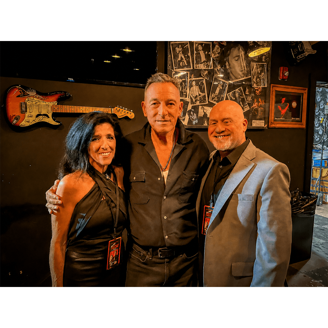 Bruce Springsteen posing with a woman and a bald man in a gray sport coat indoors against a dark wall decorated with framed photos and a mounted electric guitar.