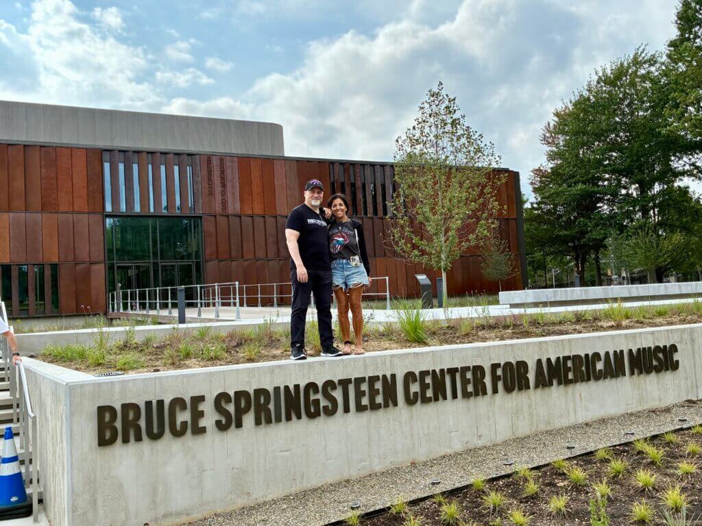Two people standing and smiling in front of the Bruce Springsteen Center for American Music building, posed behind a concrete wall displaying the center's name.