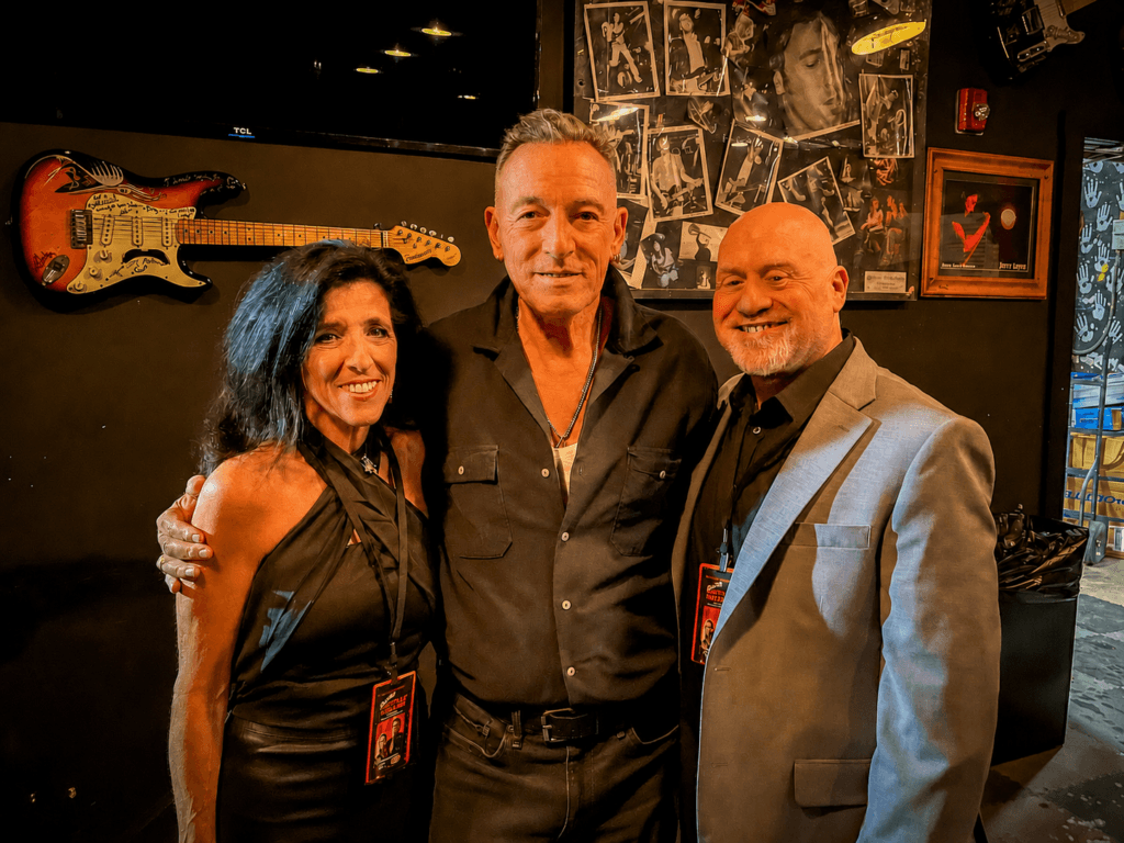 Bruce Springsteen posing with a woman and a bald man in a gray sport coat indoors against a dark wall decorated with framed photos and a mounted electric guitar.