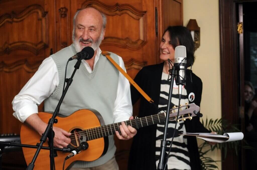 A gentleman playing an acoustic guitar while singing into a microphone next to a woman holding sheet music.