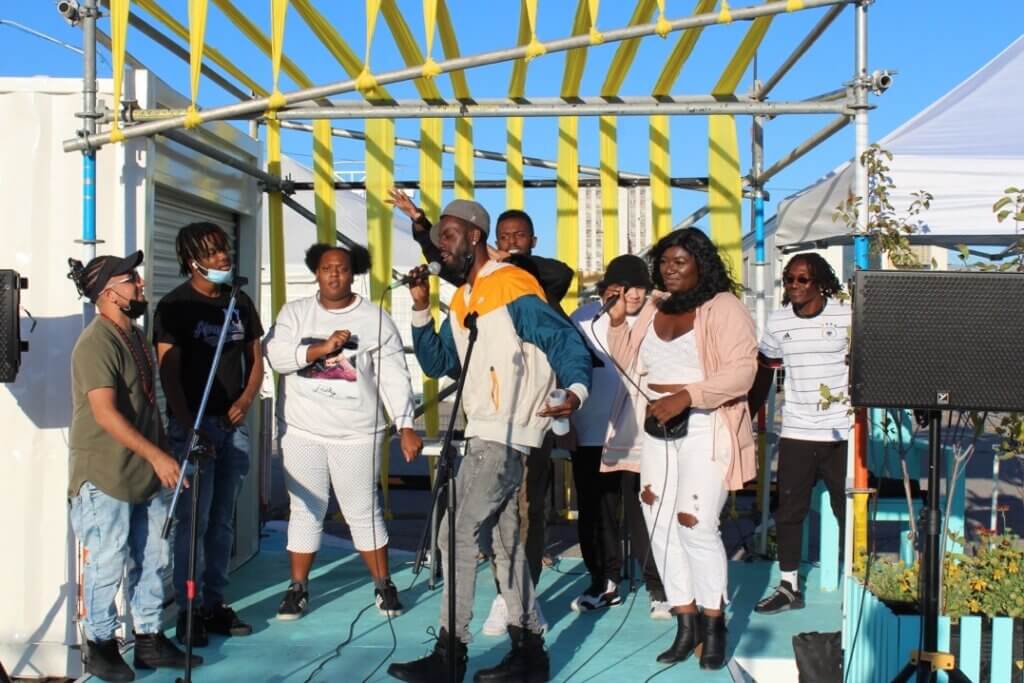 Group of people performing outdoors on a stage with yellow scaffolding and a blue floor, with one central vocalist singing into a microphone.