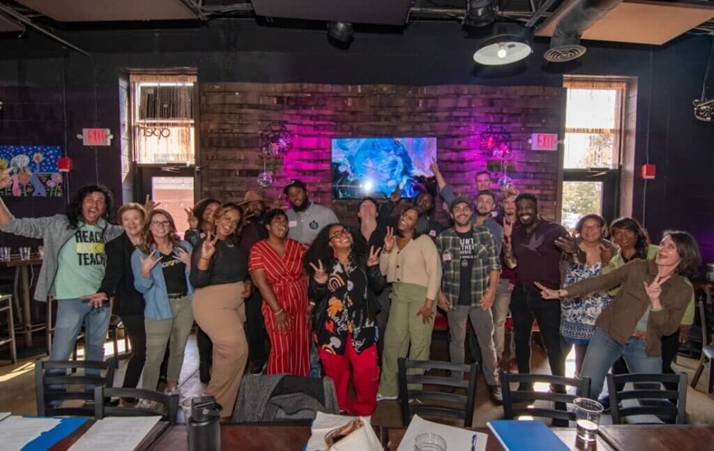 Group photo of people gathered inside an event space with a wooden accent wall, posing and smiling with tables and chairs in the foreground.