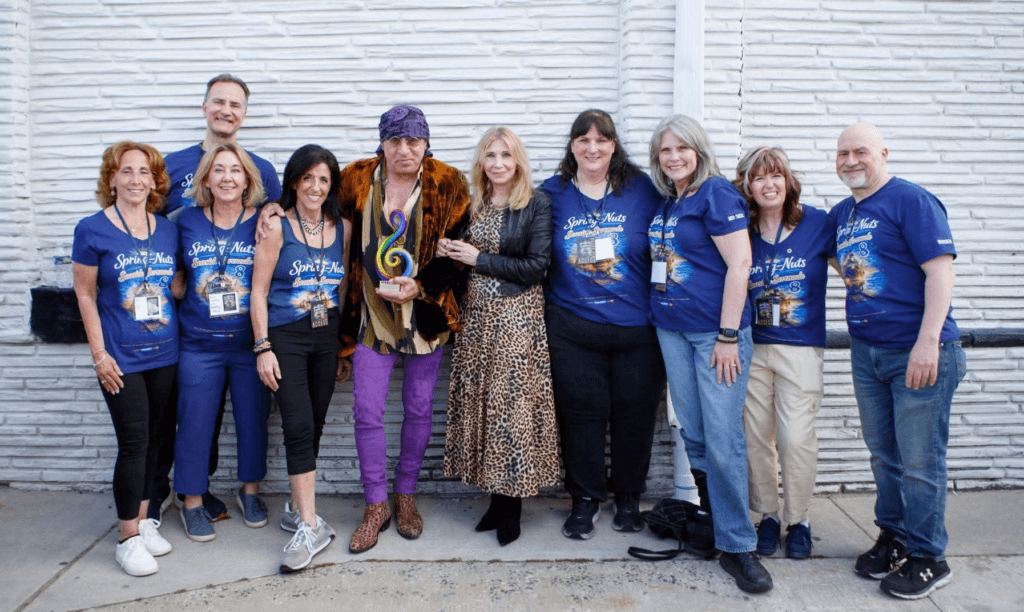 Group portrait of people posing outdoors in front of a white brick wall, with several members wearing matching blue shirts.