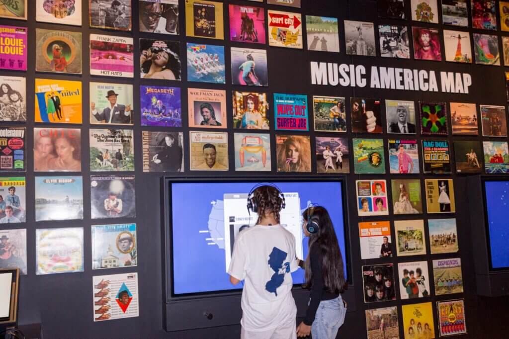 Two students wearing headphones use the Music America Map touchscreen below a wall of album covers