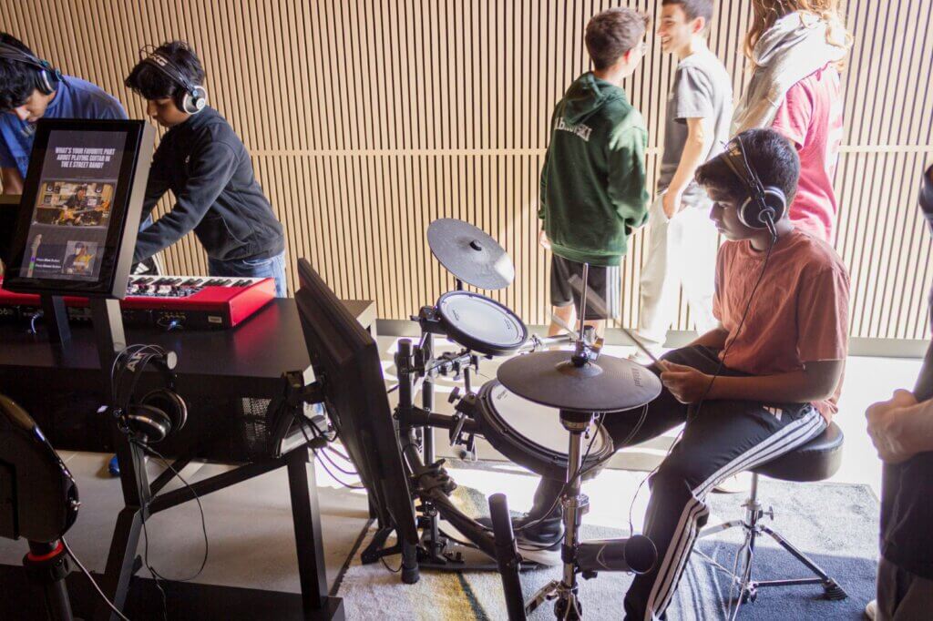 Students wearing headphones play an electronic drum kit and a keyboard at an interactive station in the Center